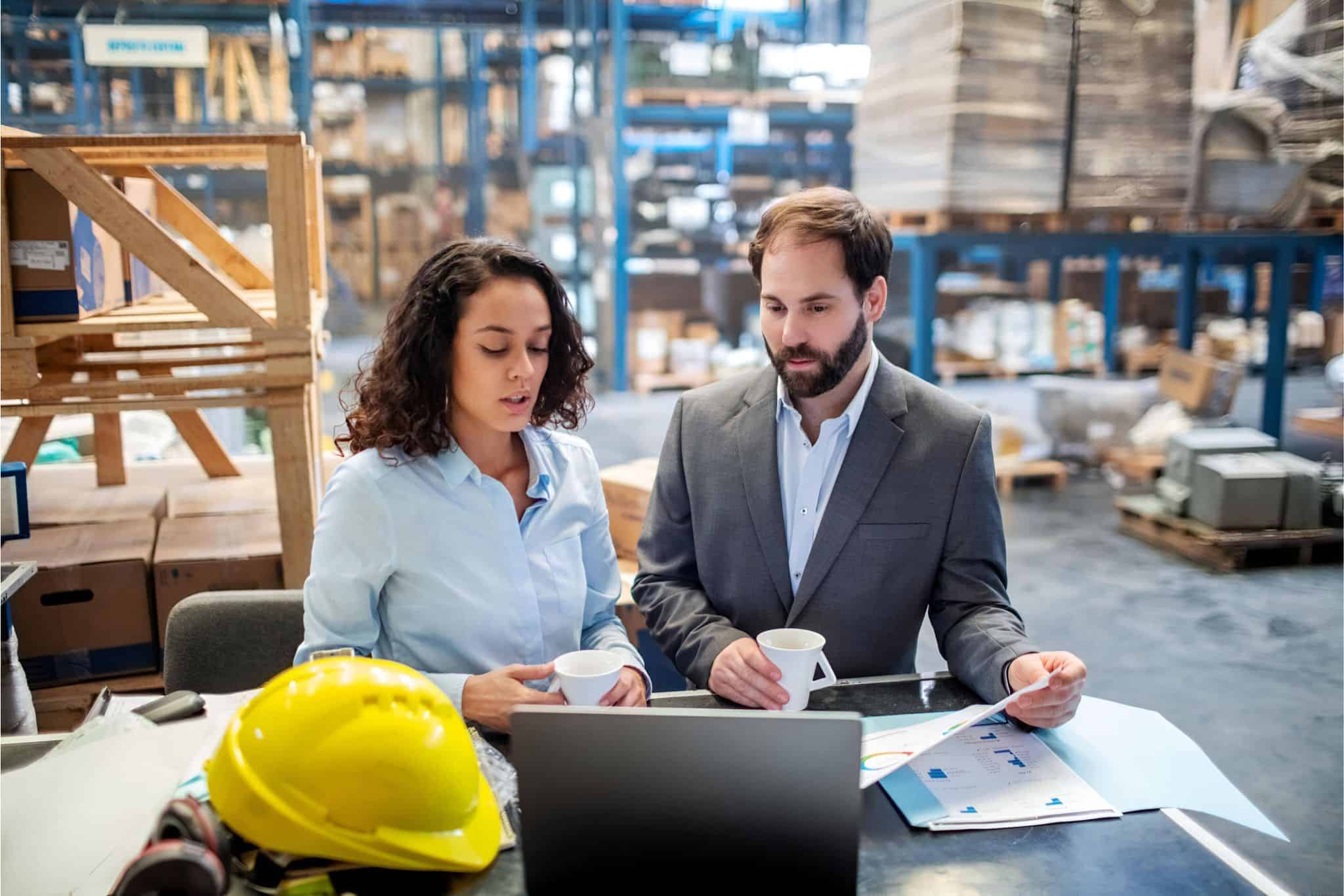 Team discussing plans on laptop in warehouse, highlighting industrial executive search with safety gear and documents nearby.