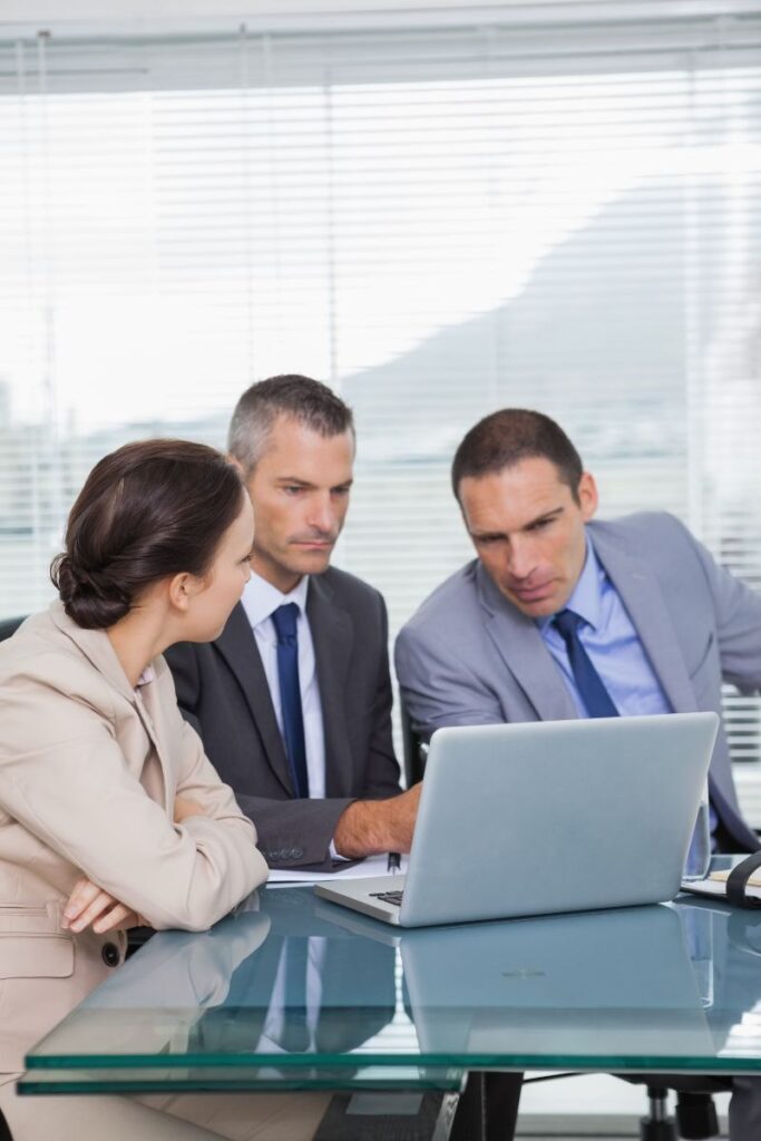 Three business professionals reviewing candidate profiles on a laptop in a corporate office, representing the services of an executive search firm.