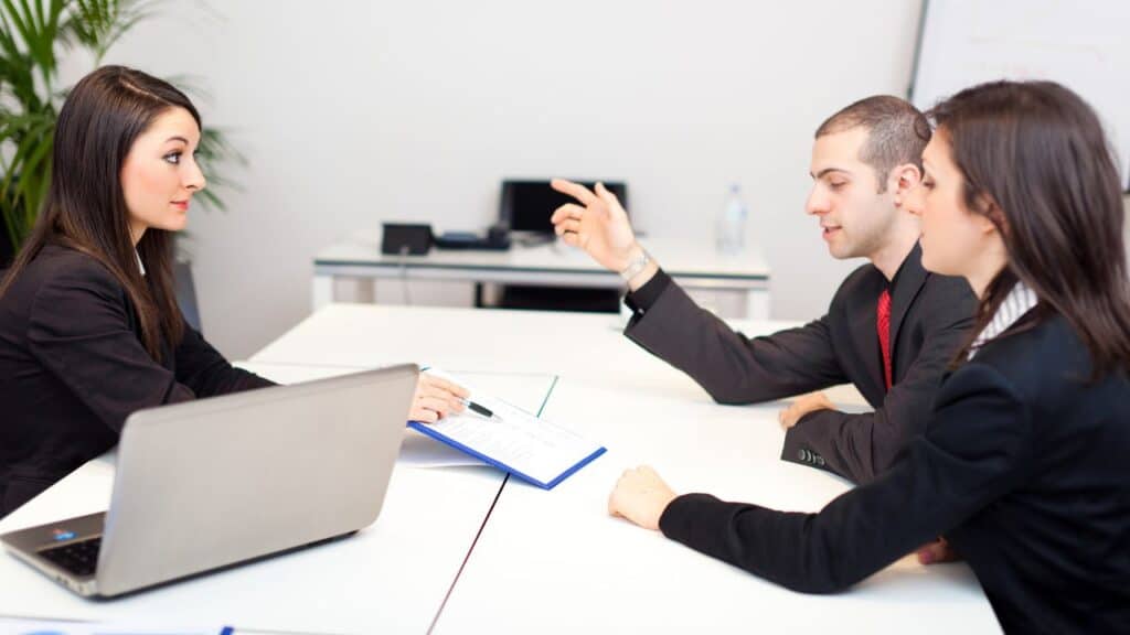 Recruiter meeting with two professionals at an executive search firm, reviewing a candidate profile in a modern office setting.