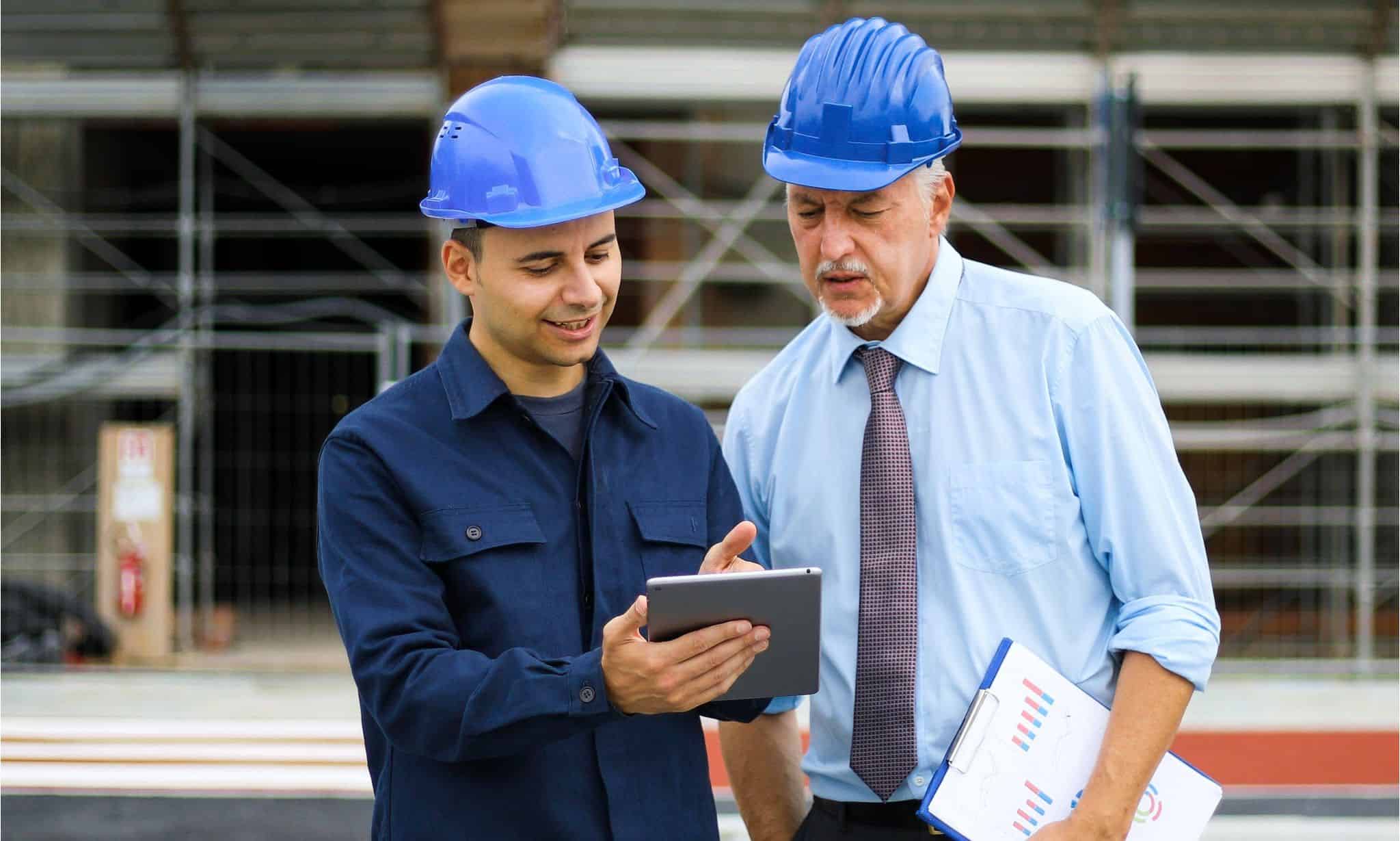 Two professionals with helmets using tablet and clipboard on construction site, representing industrial executive search expertise.