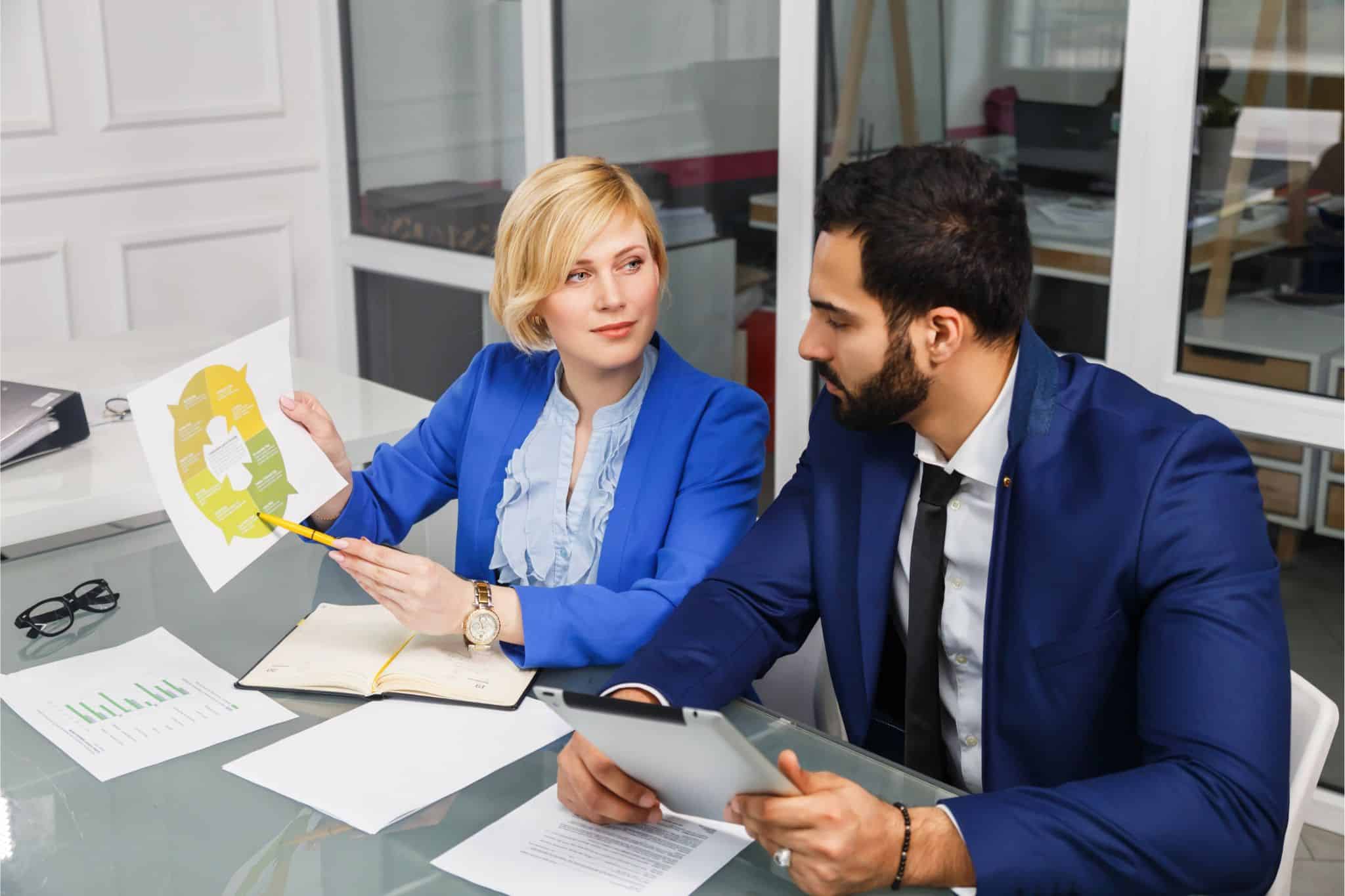 Two professionals in blue suits discussing a financial chart during an executive search meeting, with papers and a tablet on the table.